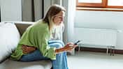 Frau mit Menstruationsbeschwerden sitzt auf dem Sofa und checkt ihre Tracking-App auf dem Smartphone