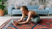 Junge Frau in Sportkleidung sitzt im Wohn-Loft auf dem Boden und schaut auf ihr Smartphone