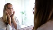 Young blond woman looking at the mirror touching her face. Female examining her facial skin looking in bathroom mirror. Wellness and lifestyle