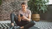 Eine Frau in Trainingskleidung sitzt auf einer Yogamatte und schaut auf ihr Handy