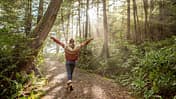 Frau läuft im Wald durch eine Waldlichtung und streckt die Arme zu den Seiten aus Frau läuft im Wald durch eine Waldlichtung und streckt die Arme zu den Seiten aus