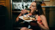 Frau sitzt in der Küche auf dem Boden mit einem Karton mit Donuts in der Hand und isst gierig einen Donut